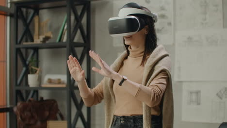 Young-woman-architect-in-office-in-virtual-reality-helmet-with-hands-commits-movements-simulating-a-graphic-interface-work.